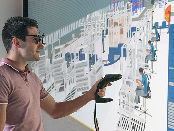 Ein junger Mann mit einer VR-Brille vor einem Bildschirm auf dem das gezeichnete Innere einer Fabrik zu sehen ist