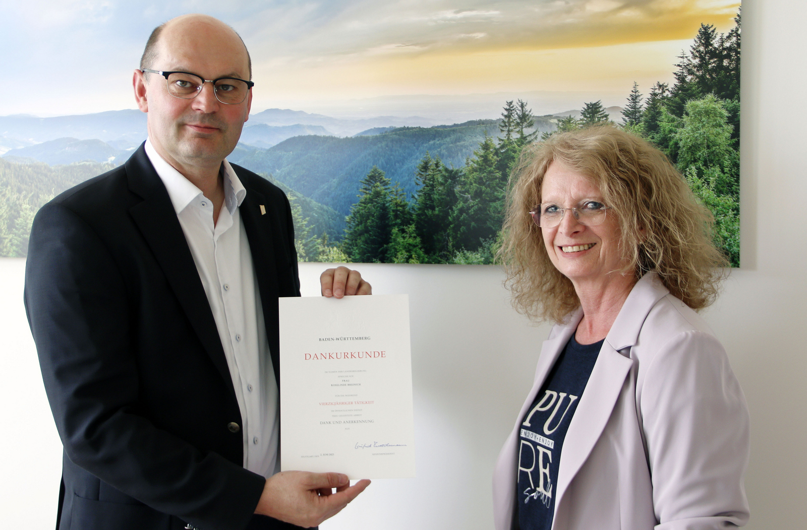 Rektor Prof. Dr. Stephan Trahasch (links) überreicht Roselinde Breinich vor einem Schwarzwaldbild die Urkunde zum Dienstjubiläum. 