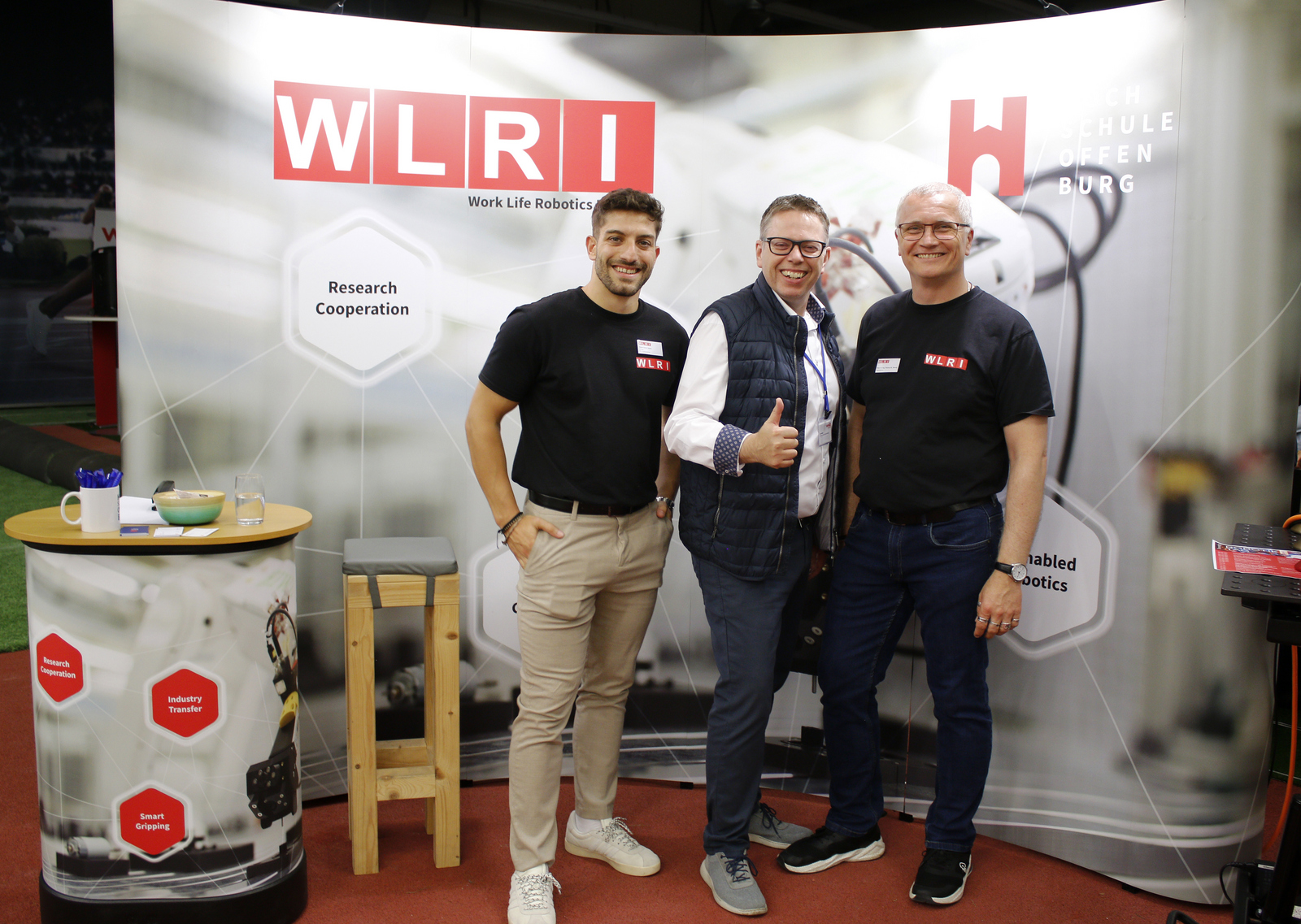 2025-07-15 wvib Robotik Tag Drei Männer stehen vor eine WLRI-WAnd, der mittlere streckt den Daumen nach oben