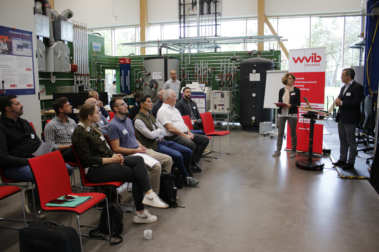 Ein Mann und eine Frau stehen vor im Technikum vor einer Heizungsanlage sitzenden Teilnehmenden
