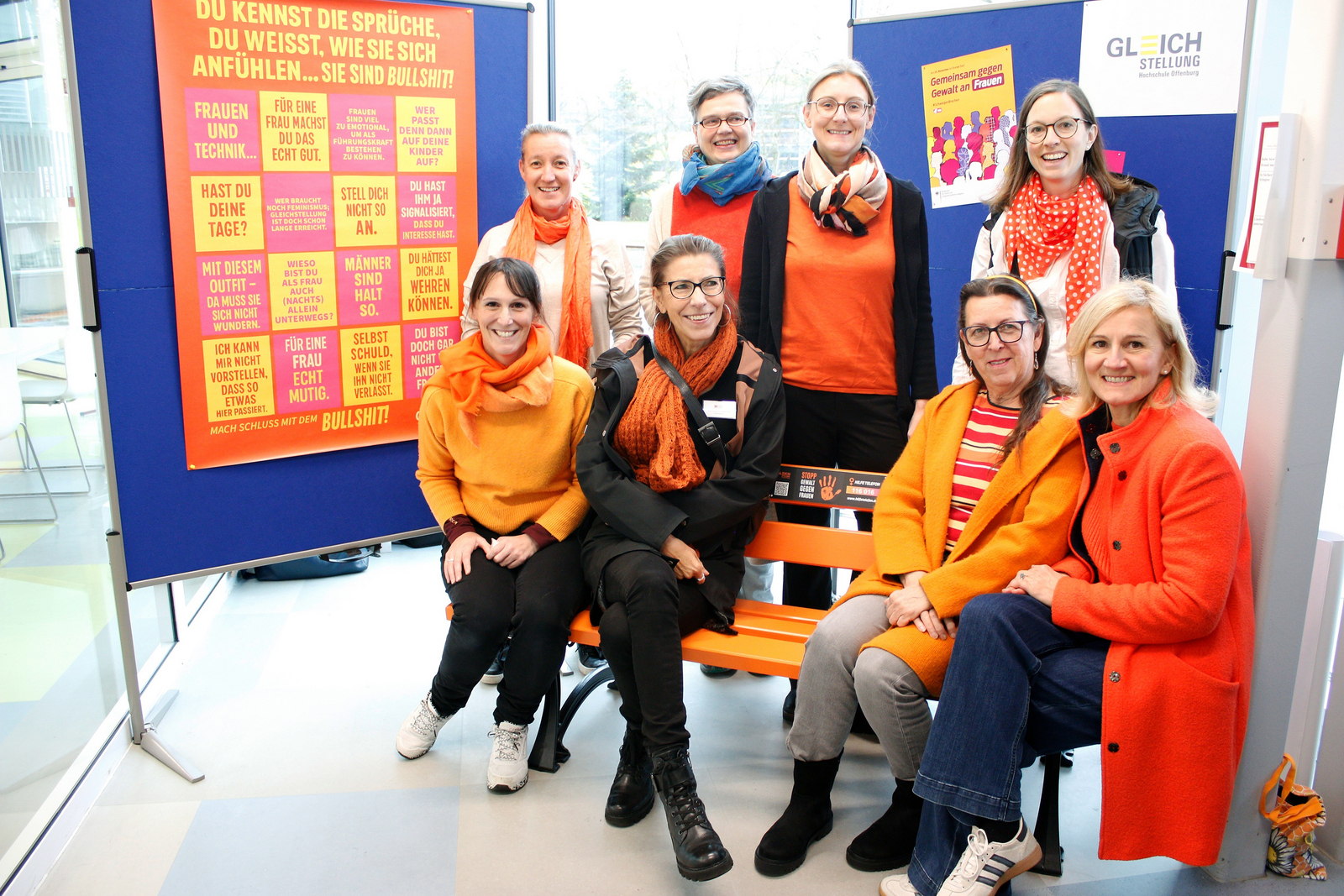 Acht Frauen mit orangenen Kleidungsstücken sitzen auf beziehungsweise stehen hinter einer orangenen Bank