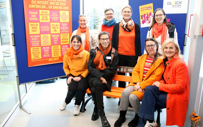 Acht Frauen mit orangenen Kleidungsstücken sitzen auf beziehungsweise stehen hinter einer orangenen Bank