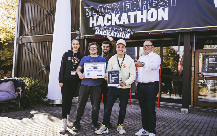 Das Gewinnerteam präsentiert vor einem Gebäude mit dem Banner Black Forest Hackathon seinen Siegerscheck und