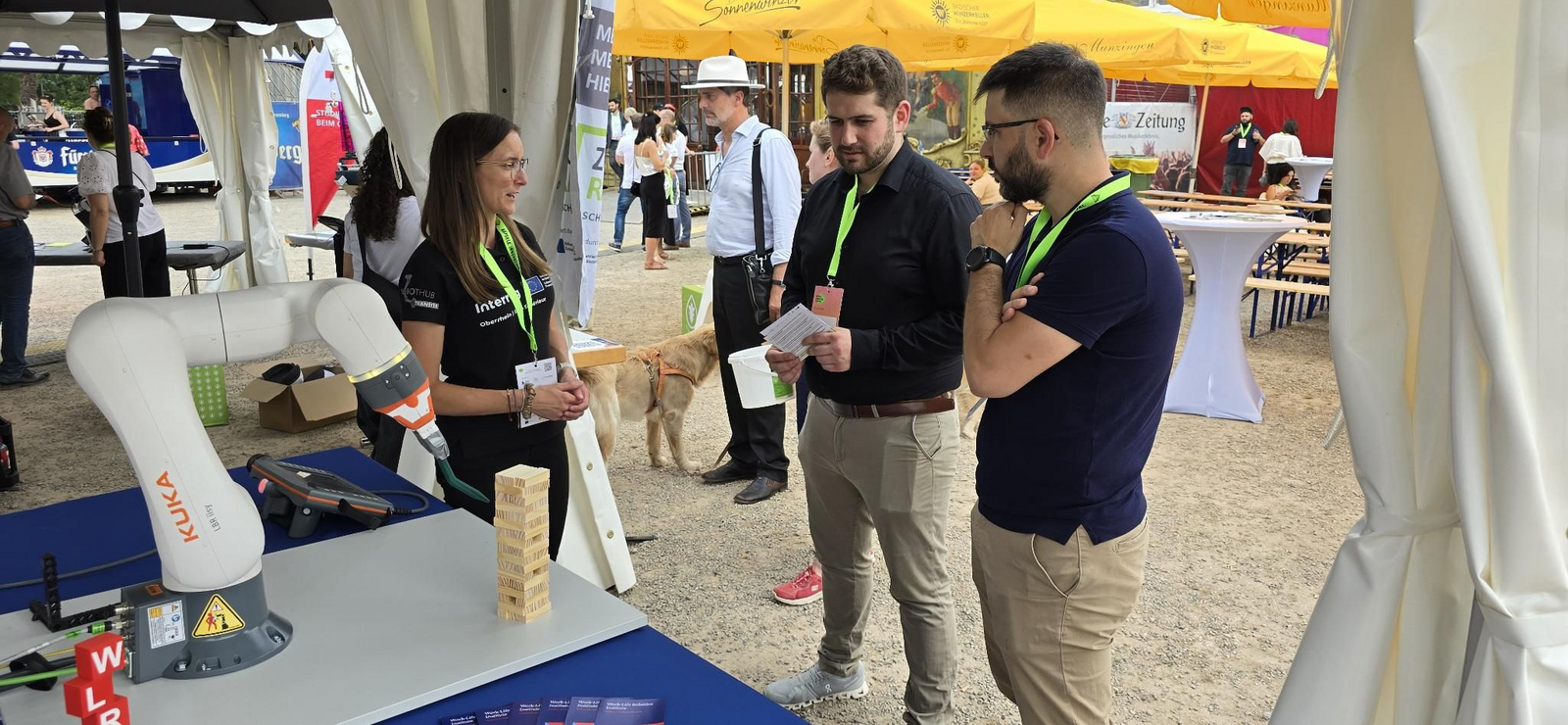 WLRI-Mitarbeiterin Jennifer Blöchle demonstriert zwei Besuchern des Innovationsfestivals einen Cobot-Arm drei Personen, die sich neben einem Roboterarm unterhalten