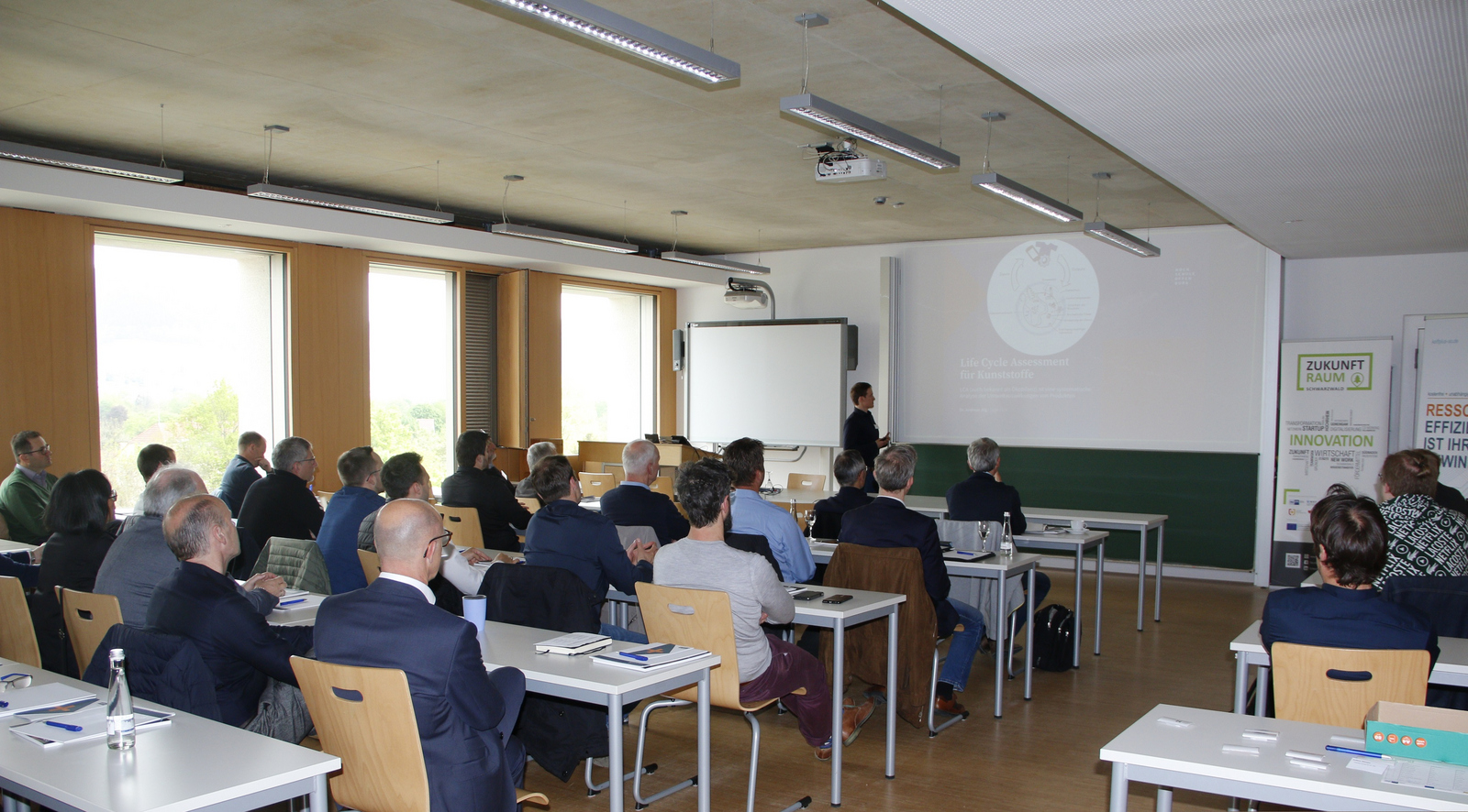 Refernt steht vor sitzenden Zuhörenden an einer Leinwand auf der eine Präsentation zu sehen ist