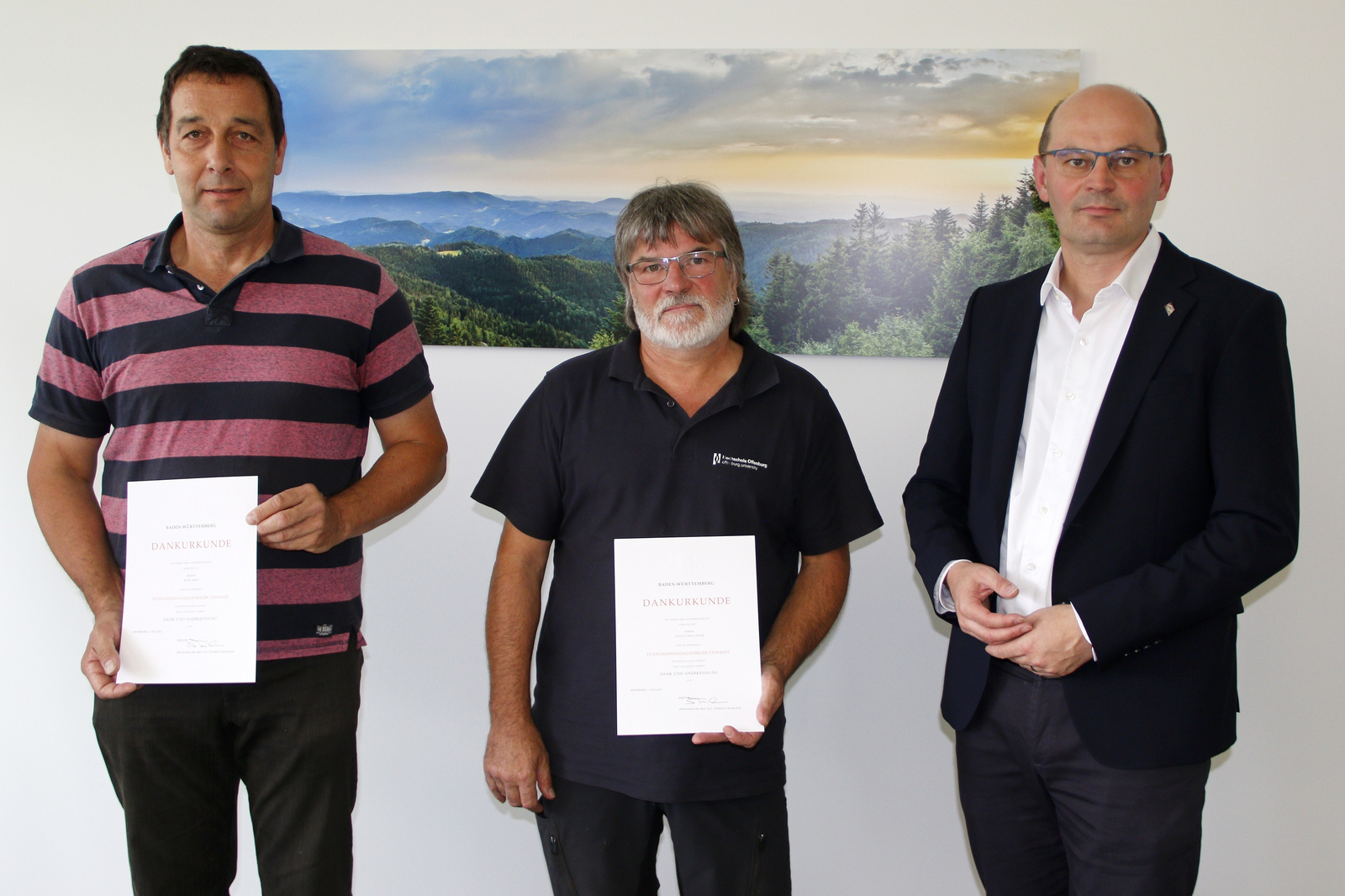 Rudi Kirn (links) und Stefan Hollinger (Mitte) stehen im Büro des Rektors Stephan Trahasch (rechts) vor einem Bild mit einer Schwarzwaldlandschaft.