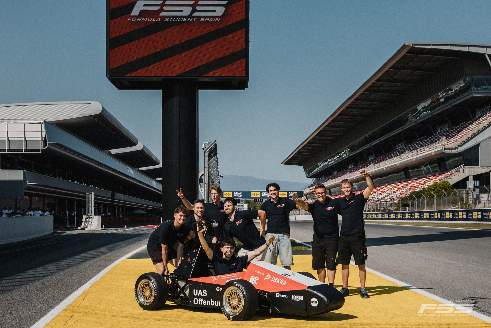 2025-08-15 Web-Meldung BFFT sammelt wertvolles Wissen Das Team in lustigen Posen hinter dem Auto auf dem Circuit.