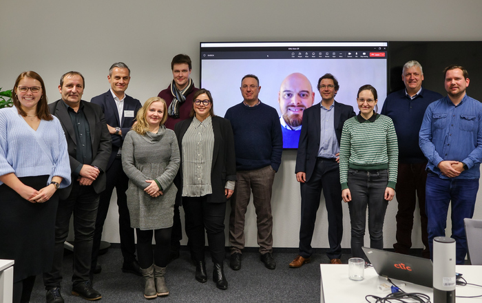 Gruppenbild der Projektbeteiligten vor einem Bildschirm auf dem ein online zugeschalteter Projektbeteiligter zu sehen ist