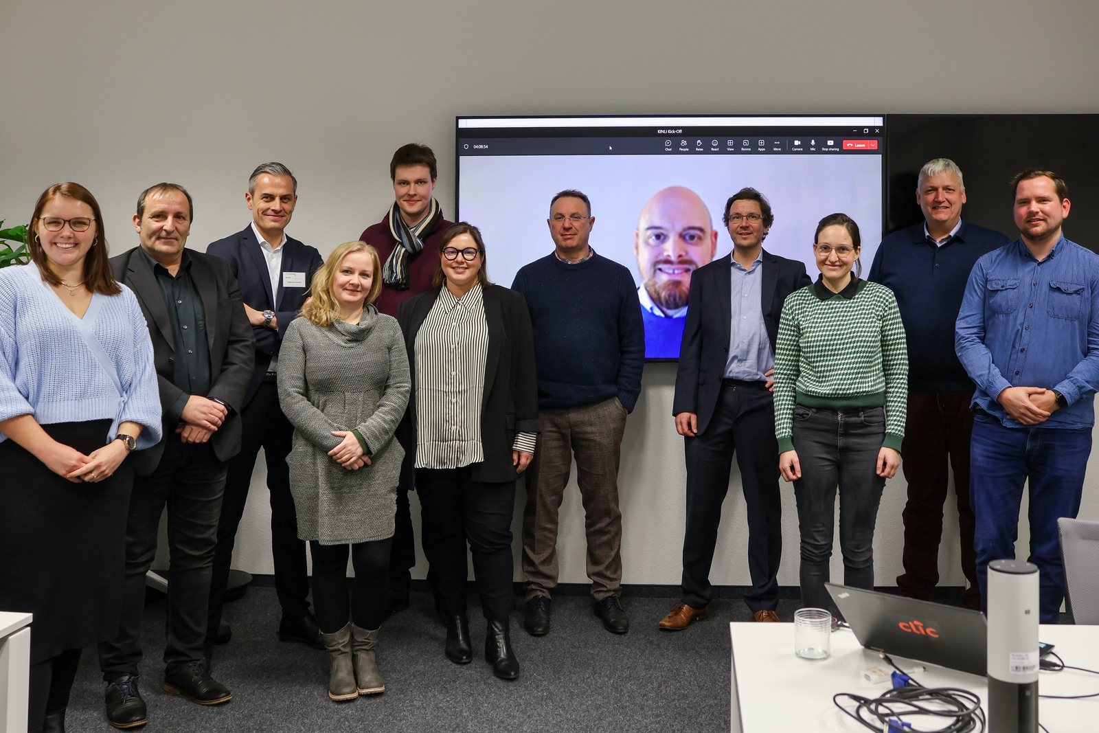 Gruppenbild der Projektbeteiligten vor einem Bildschirm auf dem ein online zugeschalteter Projektbeteiligter zu sehen ist