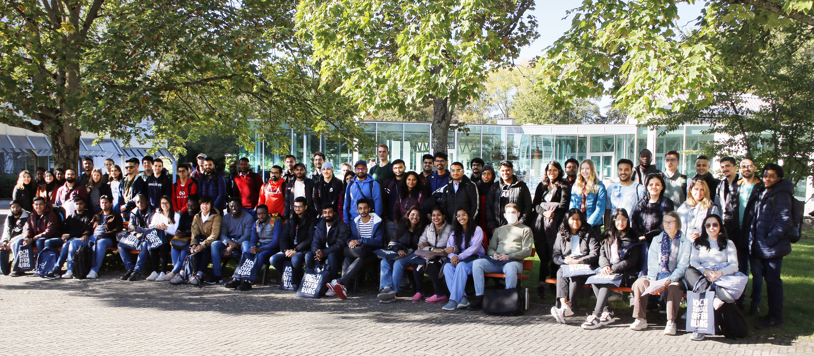 Die neuen internationalen Studierenden stehen beziehungsweise itzen unter Bäumen auf dem Campus