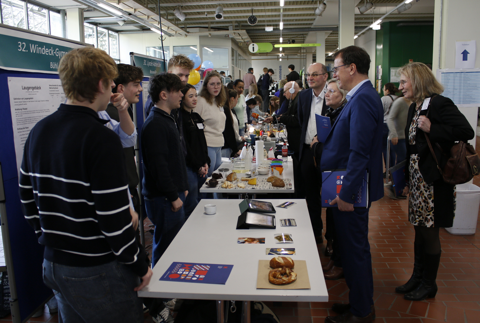 Mehrere Erwachsene stehen vor einem Tisch mit Laugengebäck und lassen sich von Schülerinnen und Schülern etwas erklären.