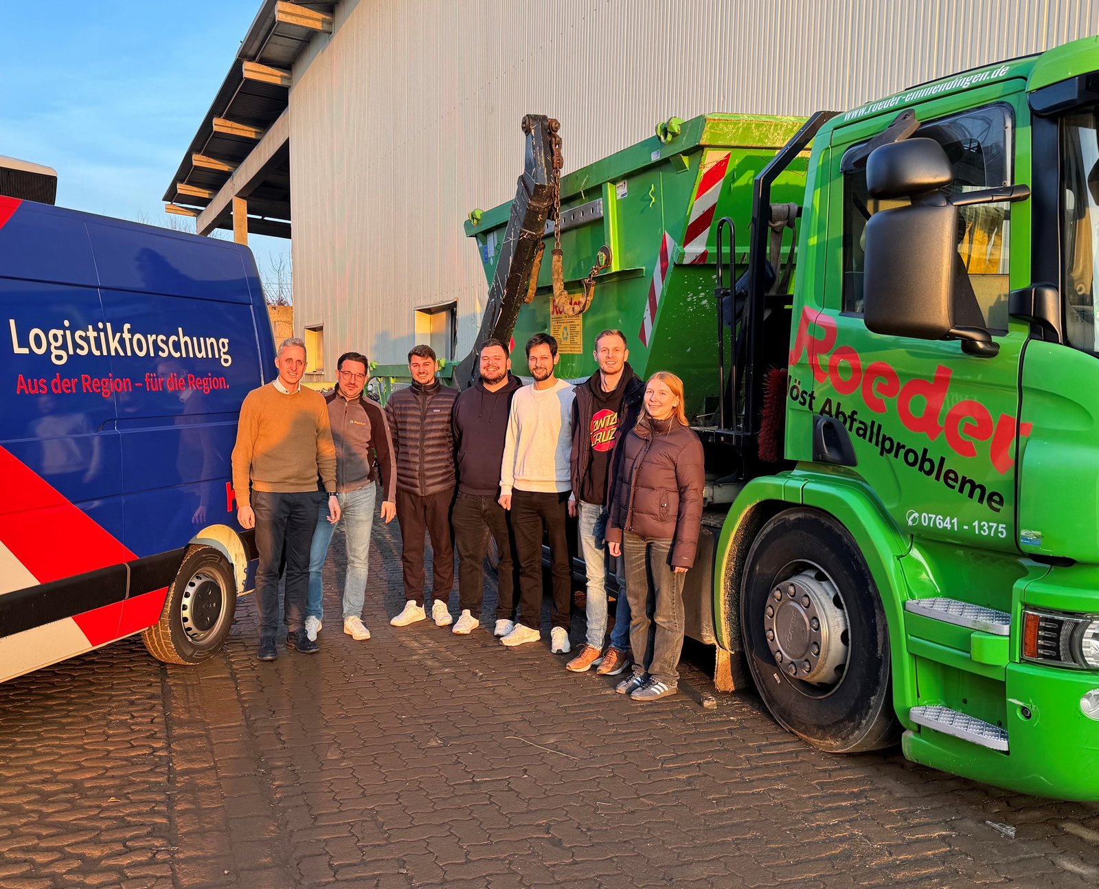 Eine Gruppe von sieben Personen steht zwischen einem blauen Transporter mit der Aufschrift 'Logistikforschung. Aus der Region – für die Region' und einem grünen Lkw mit der Aufschrift 'Roeder löst Abfallprobleme' vor einer Lagerhalle.