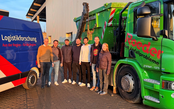 Eine Gruppe von sieben Personen steht zwischen einem blauen Transporter mit der Aufschrift 'Logistikforschung. Aus der Region – für die Region' und einem grünen Lkw mit der Aufschrift 'Roeder löst Abfallprobleme' vor einer Lagerhalle.