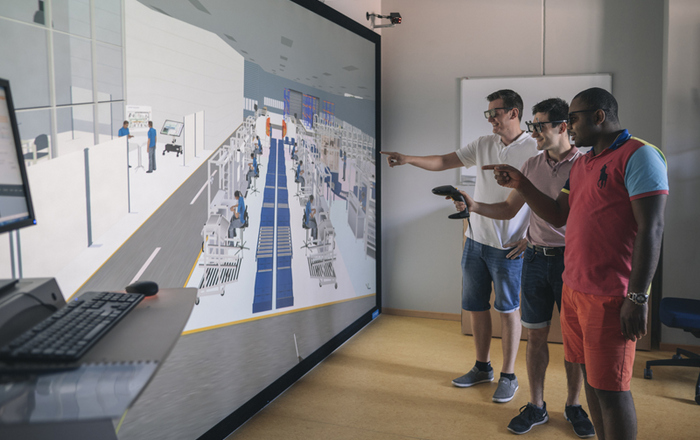 drei Personen stehen vor einem großen Bildschirm auf dem eine virtuelle Fabrik zu sehen ist
