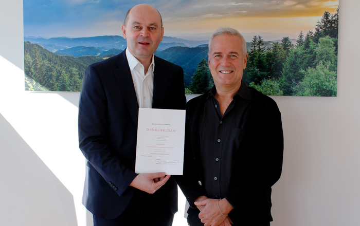 Zwei Männer im Anzug vor einem Schwarzwaldfoto halten eine Urkunde zwischen sich