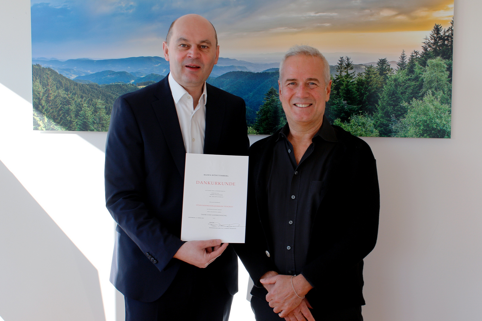 Zwei Männer im Anzug vor einem Schwarzwaldfoto halten eine Urkunde zwischen sich