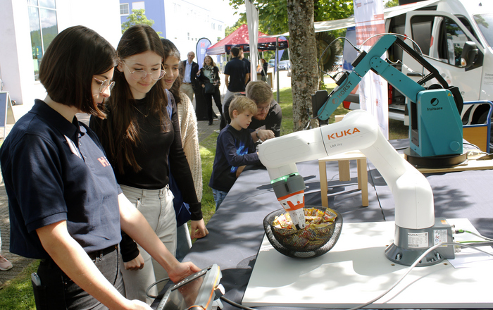 jungen menschen stehen an einem Tisch auf dem zwei Robotergreifarme aufgebuat sind und versuchen Klötzchen zu stapeln beziehungsweise Süßigkeiten zu greifen