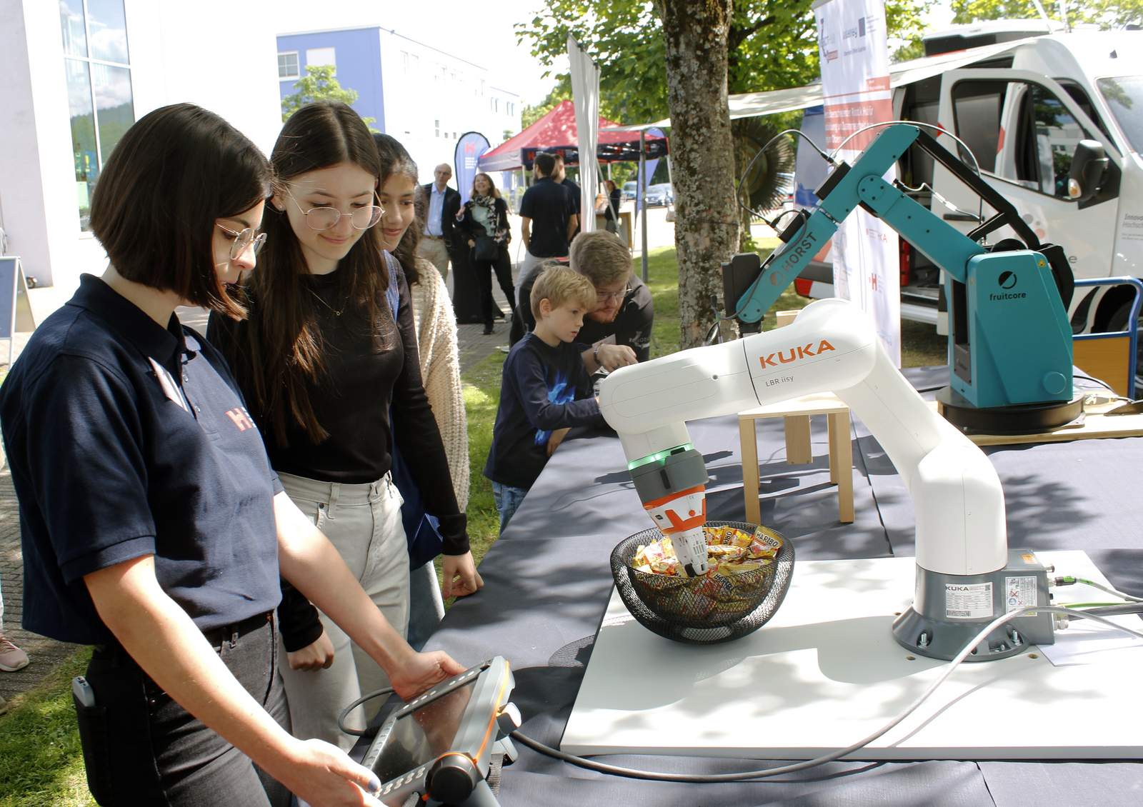 jungen menschen stehen an einem Tisch auf dem zwei Robotergreifarme aufgebuat sind und versuchen Klötzchen zu stapeln beziehungsweise Süßigkeiten zu greifen