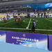 Auf einem Fuballfeld in einer Halle spielen drei rotgkleidete Roboter gegen drei blaugekleidete, von denen einer gerade eine Freistoß an der Seitenlinie ausführt. Am Spielfeldrand stehen Menschen und auf den Tribünen sitzen zahlreiche Zuschauende.
