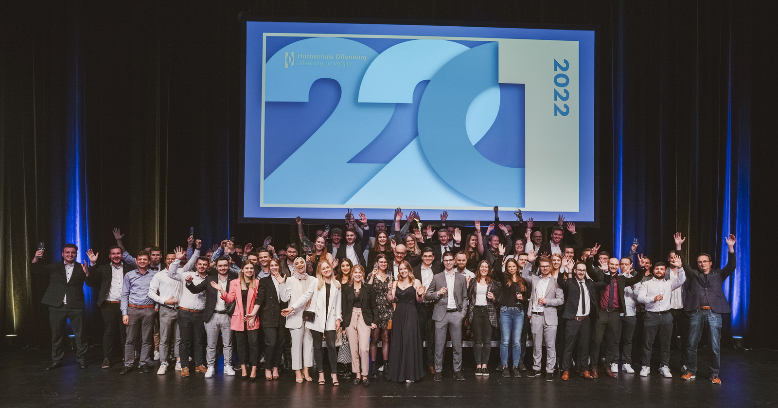 Gruppenfoto der jubelnden Absolvent*innen unter dem Logo der Abschlussfeier