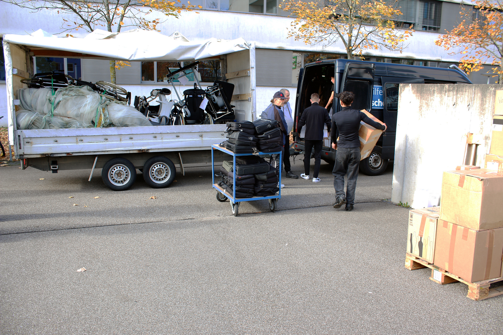 Mehrere Männer packen Kisten und Kaptops in einen Kastenwagen mit Anhänger auf dem Rollstühle und Netze aus dem Obst- und Weinbau liegen