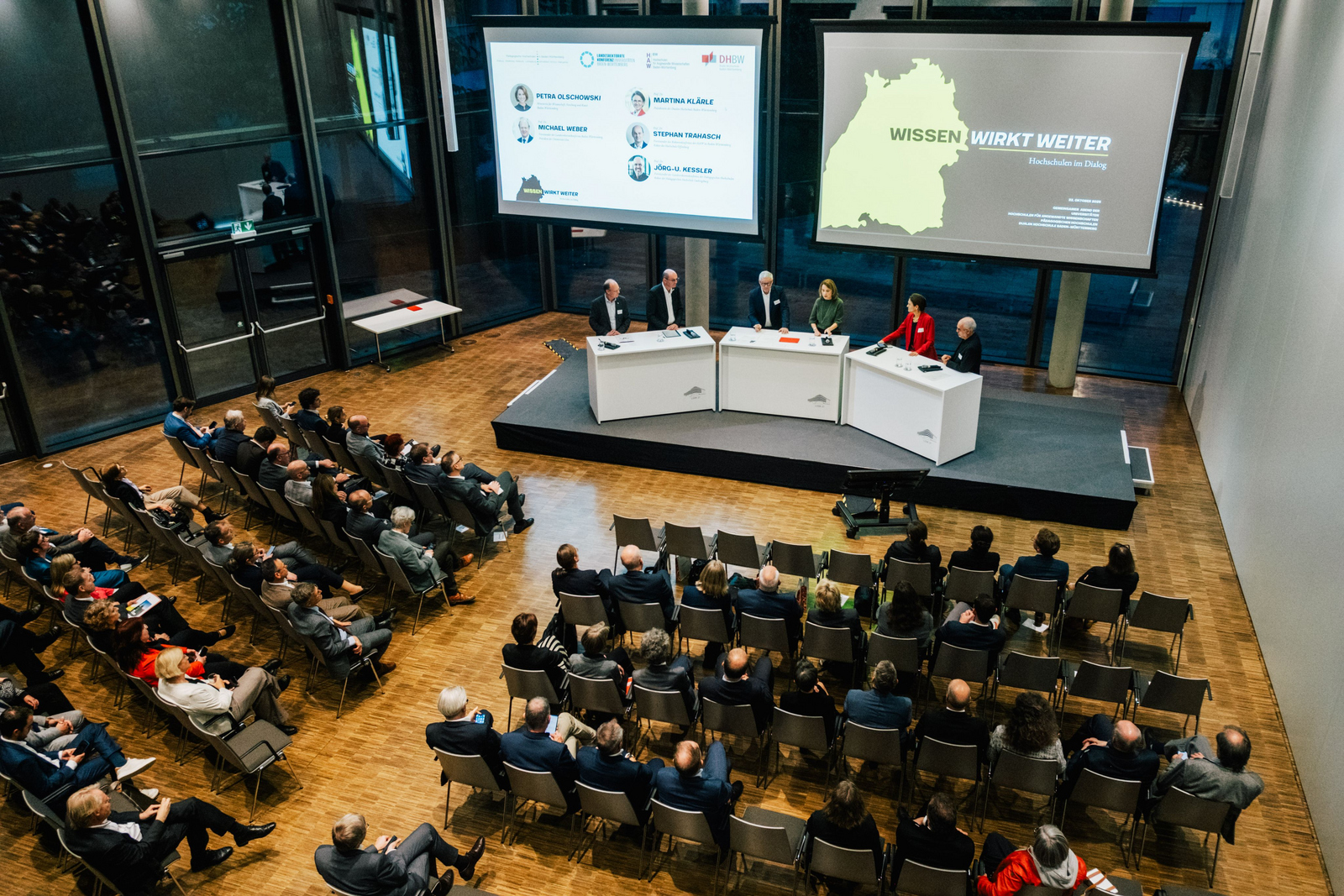 2025-10-27 Web-Meldung Hochschulen im Dialog Mehrere Menschen stehen auf einer Bühne im Halbkreis an Rednerpulten hinter ihne zwei große Bidlschirme mit Präsentationen und vor ihnen sitzende Zuhörende