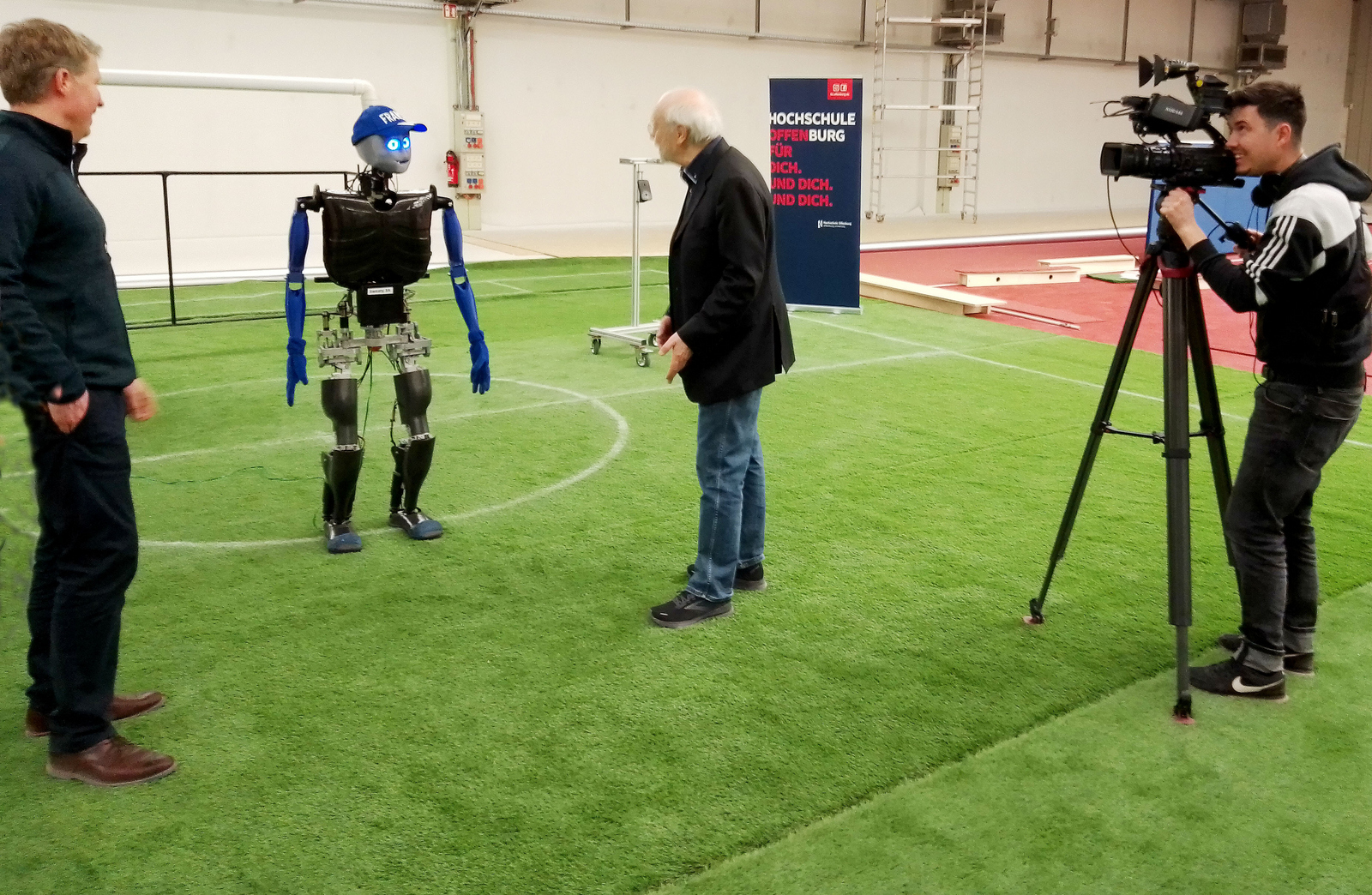 Prof. Hensel, Sweaty und Prof. Hochberg stehen auf einem Fußballfeld, unterhalten sich und werden dabei von einem Kameramann gefilmt  