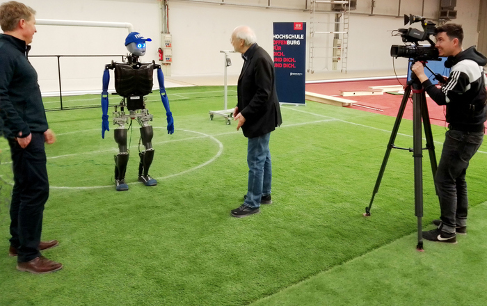 Prof. Hensel, Sweaty und Prof. Hochberg stehen auf einem Fußballfeld, unterhalten sich und werden dabei von einem Kameramann gefilmt  