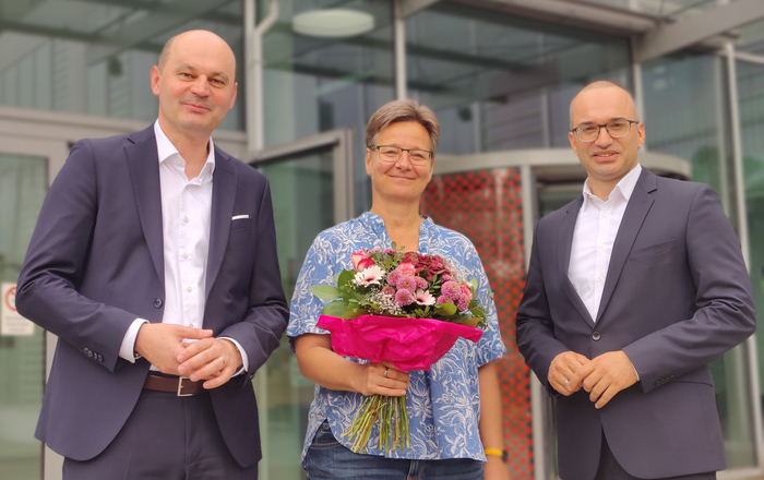 Die drei Personen stehn vor Gebäude D, Grit Köhler hält einen Blumenstrauß in der Hand