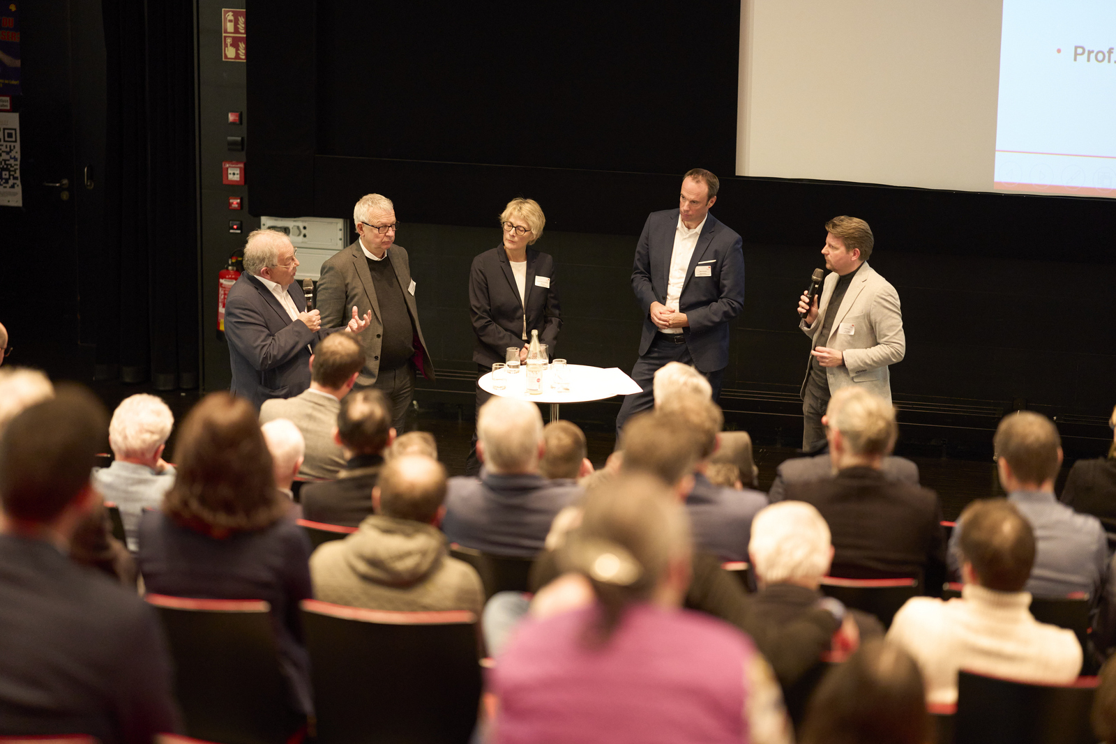 Fünf Personen stehen in einem Hörsaal vor sitzenden Zuhörenden um einen Stehtisch herum und diskutieren