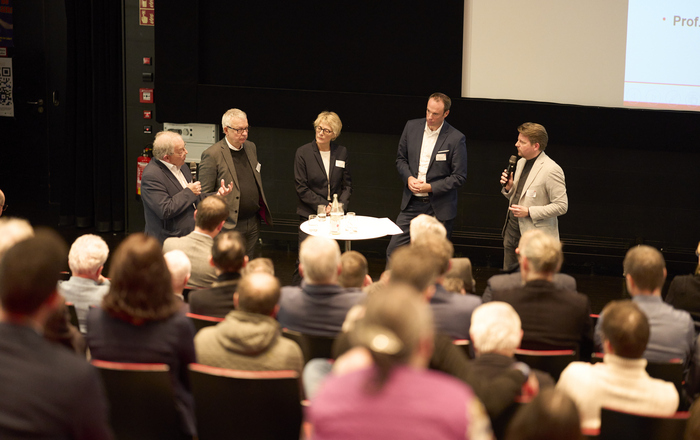 Fünf Personen stehen in einem Hörsaal vor sitzenden Zuhörenden um einen Stehtisch herum und diskutieren