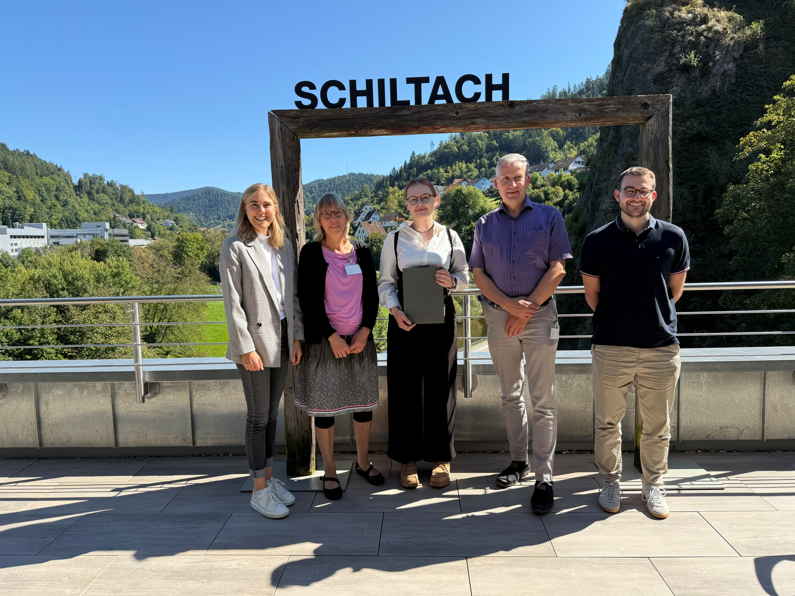 Fünf Menschen stehen bei strahlendem Sonnenschein vor einem großen Holzrahmen auf dem Schiltach steht und der eine Aussicht auf den gleichnamigen Ort einrahmt.