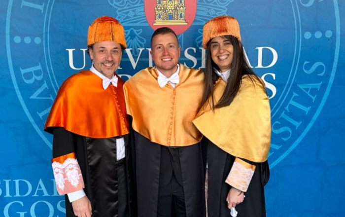 Zwei Männer und eine Frau Arm in Arm in Doktorroben vor dem Wappen der Universidad de Burgos