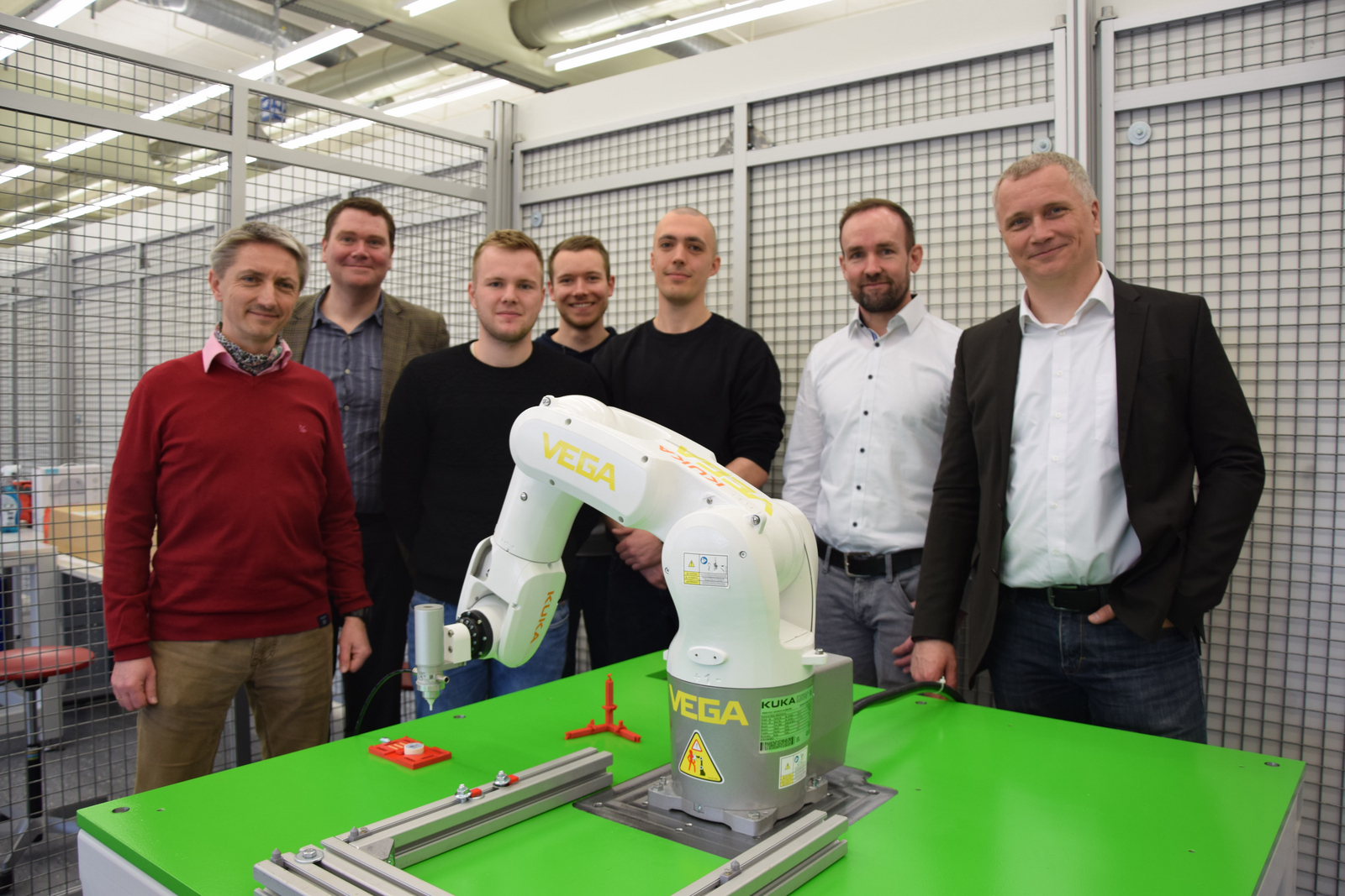 Die Projektbeteiligten stehen hinter dem Tisch mit dem Roboter der programmiert wird.