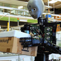 Ein humanoider Roboter steht vor einer Maschine und hält ein Paket vor einen Bildschirm.  