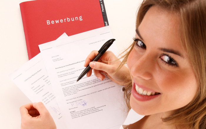Eine junge Frau schreibt an ihre Bewerbung, eine rote Bewerbungsmappe liegt auf dem Tisch