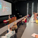 Eine Frau steht vor einer großen Leinwand und spricht zu sitzenden Zuhörenden in einem Hörsaal