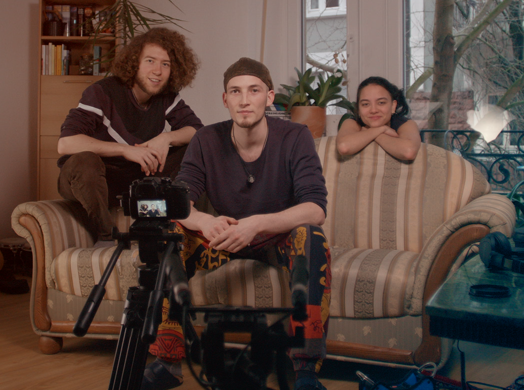 Die drei sitzen in einem Wohnzimmer auf beziehungsweise neben einem Sofa und filmen sich selbst.