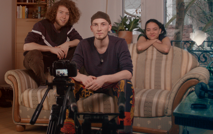 Die drei sitzen in einem Wohnzimmer auf beziehungsweise neben einem Sofa und filmen sich selbst.