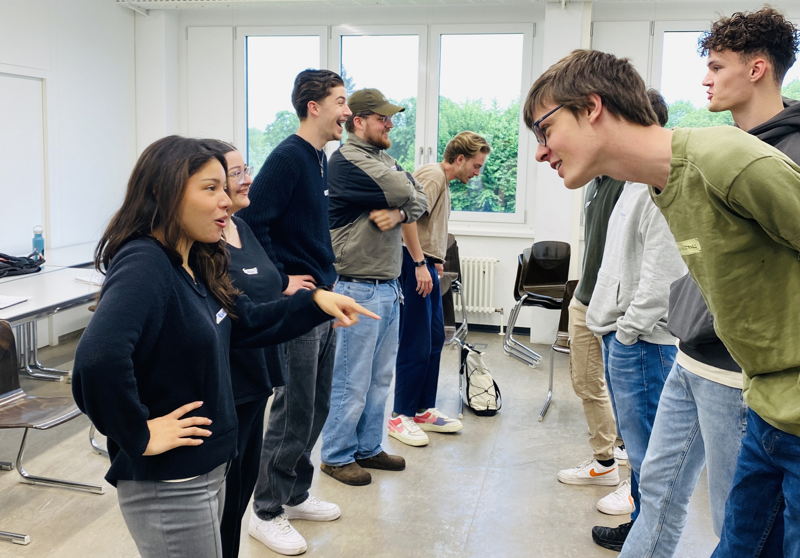 Gruppe aus Studierenden bei einem Workshop in einem Raum. In einer Reihe stehen sich immer 2 Personen gegenüber und interagieren miteinander. Im Hintergrund sind Fenster zu sehen.