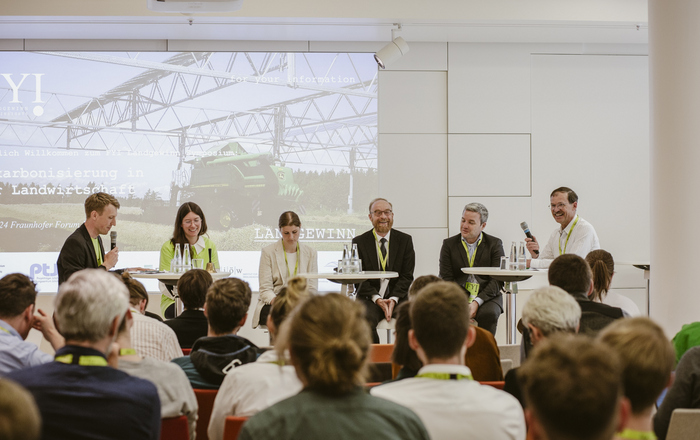 Die Beteiligten der Podiumsdiskussion sitzen nebeneinander auf der bühne vor den sitzenden Zuhörenden