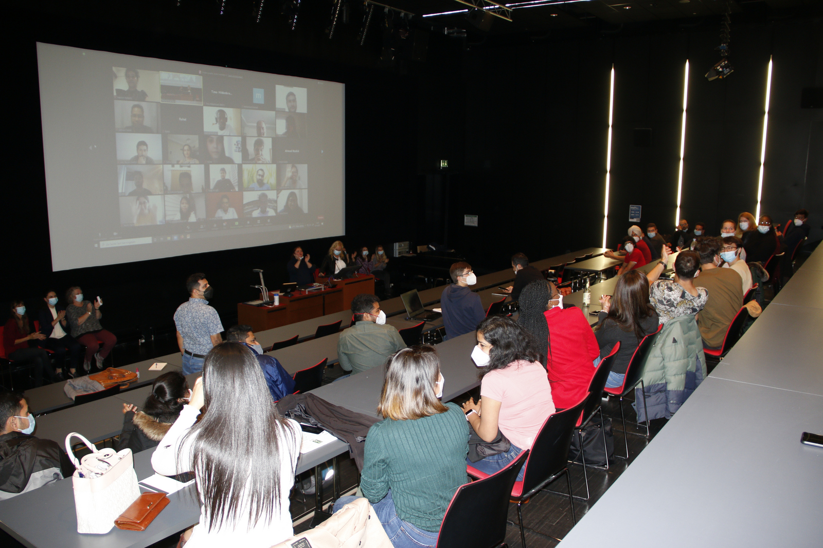 Das Bild zeigt Internationale Studierende in einem Hörsaal und auf einer Leinwand die Gesichter der noch in ihren Heimatländern weilenden in Form von Zoom-Kacheln. 