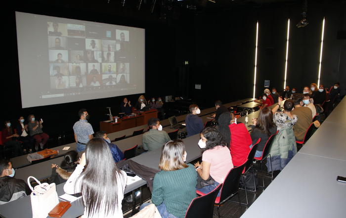 Das Bild zeigt Internationale Studierende in einem Hörsaal und auf einer Leinwand die Gesichter der noch in ihren Heimatländern weilenden in Form von Zoom-Kacheln. 