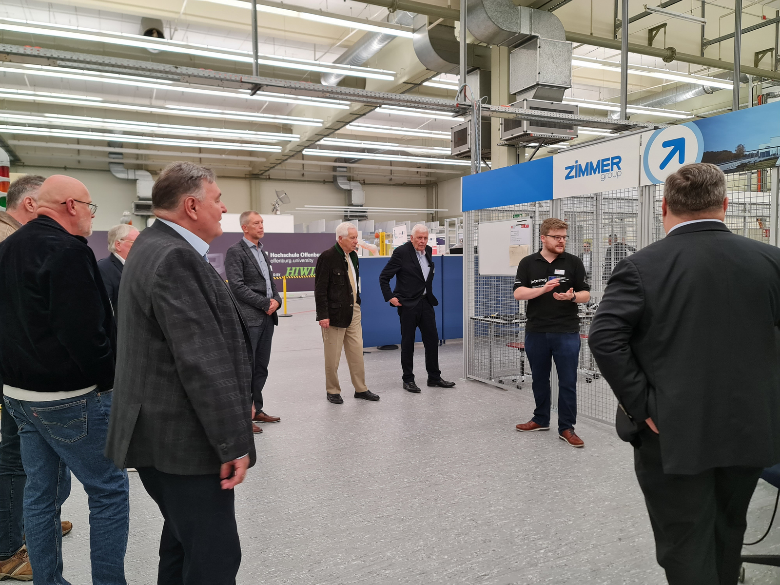Menschen stehen im HAlbkreis in einer Industriehalle vor einem Metallkäfig, eienr eklärt etwas 
