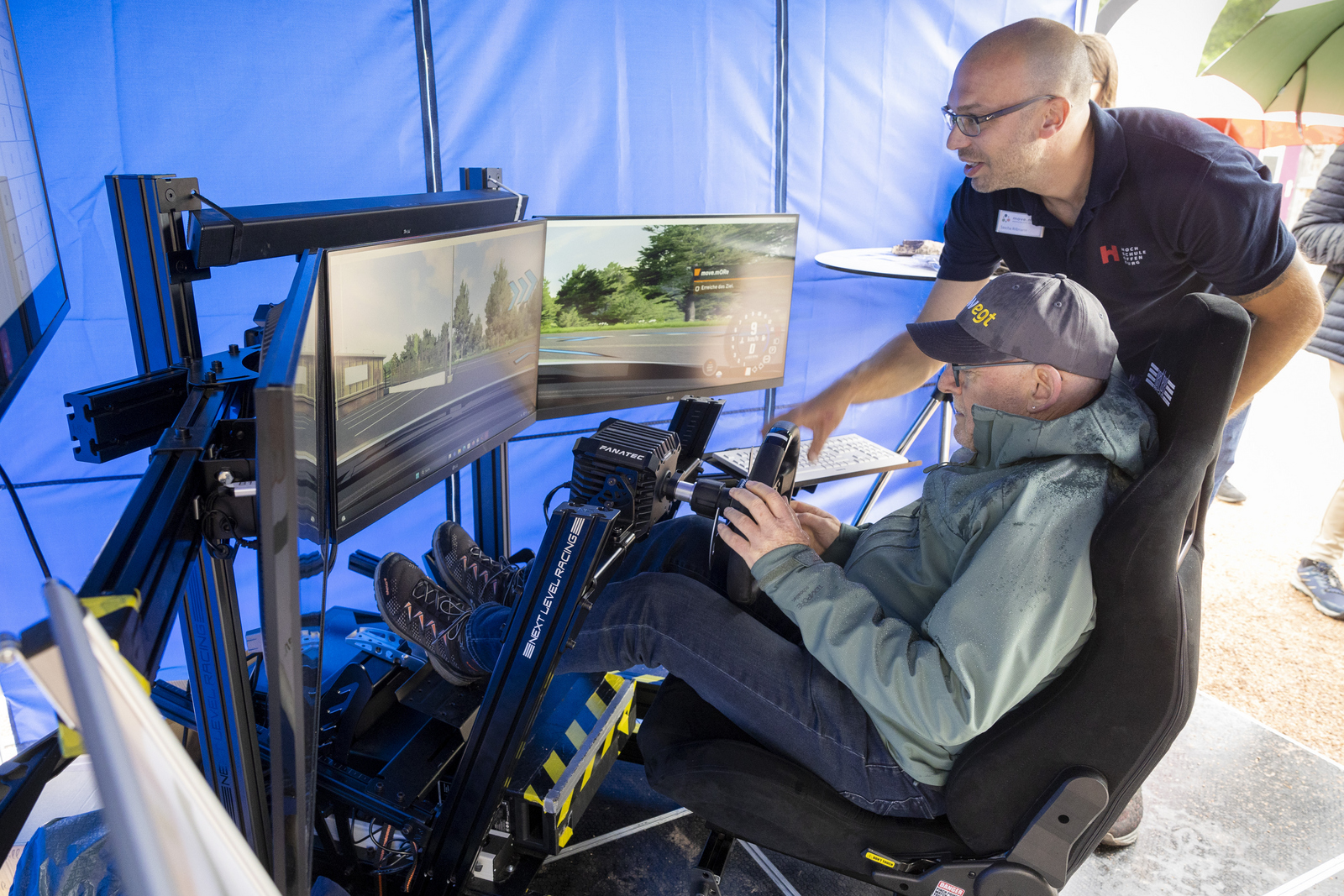Mann im E-Auto-Fahrsimulator, ein anderer Mann steht daneben und erklärt etwas