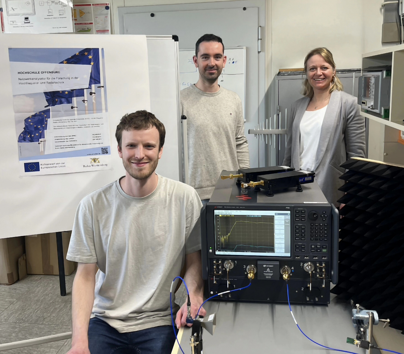 zwei Männer und eine Frau sitzen beziehunsgweise stehen um den Netzwerkanalysator, der auf einem Tisch steht, herum