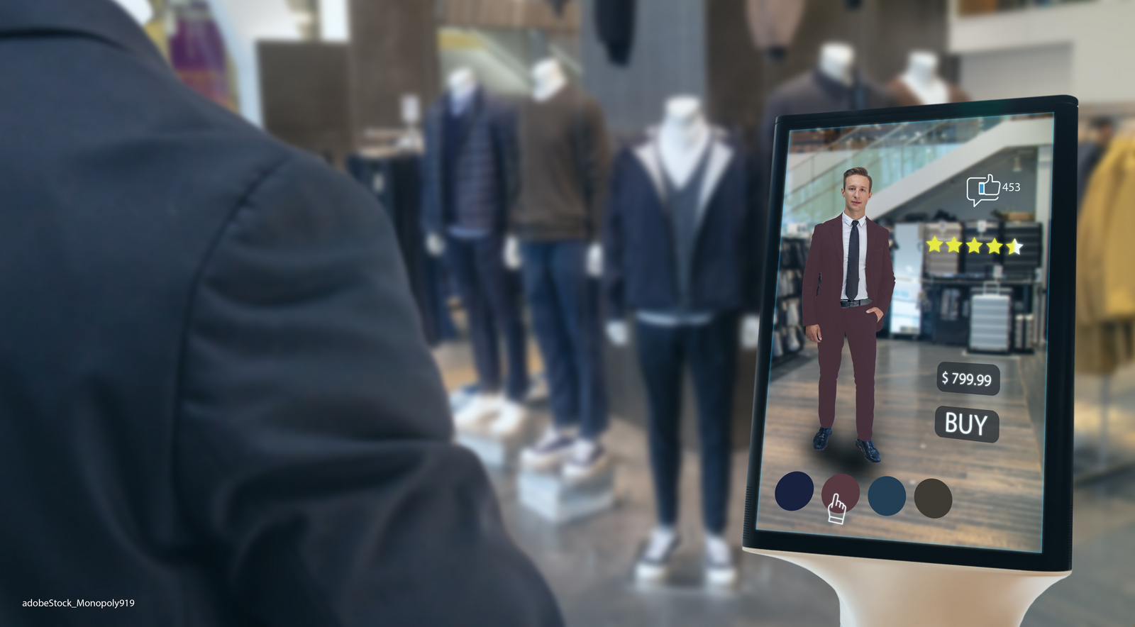 In einem Herrengeschäft stehen Schaufensterpuppen mit verschiedenen Kleidungsstücken, auf einem Tabletbildschirm ist zu sehen wie eins der Kleidungsstücke an einem Mann aussehen würde.