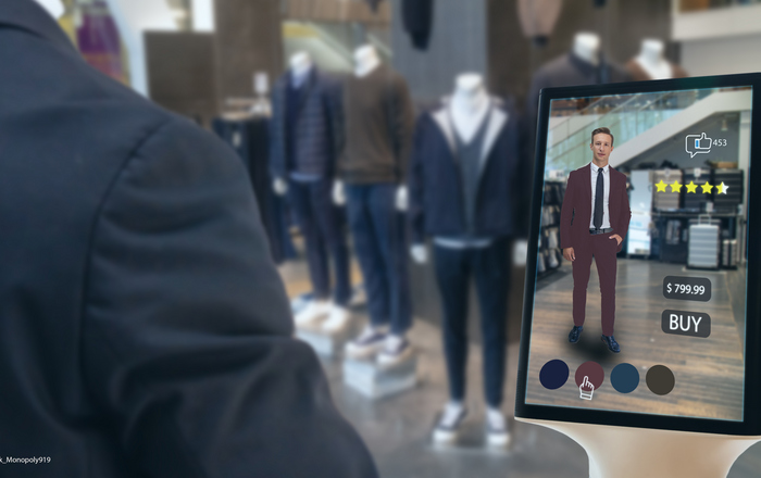 In einem Herrengeschäft stehen Schaufensterpuppen mit verschiedenen Kleidungsstücken, auf einem Tabletbildschirm ist zu sehen wie eins der Kleidungsstücke an einem Mann aussehen würde.