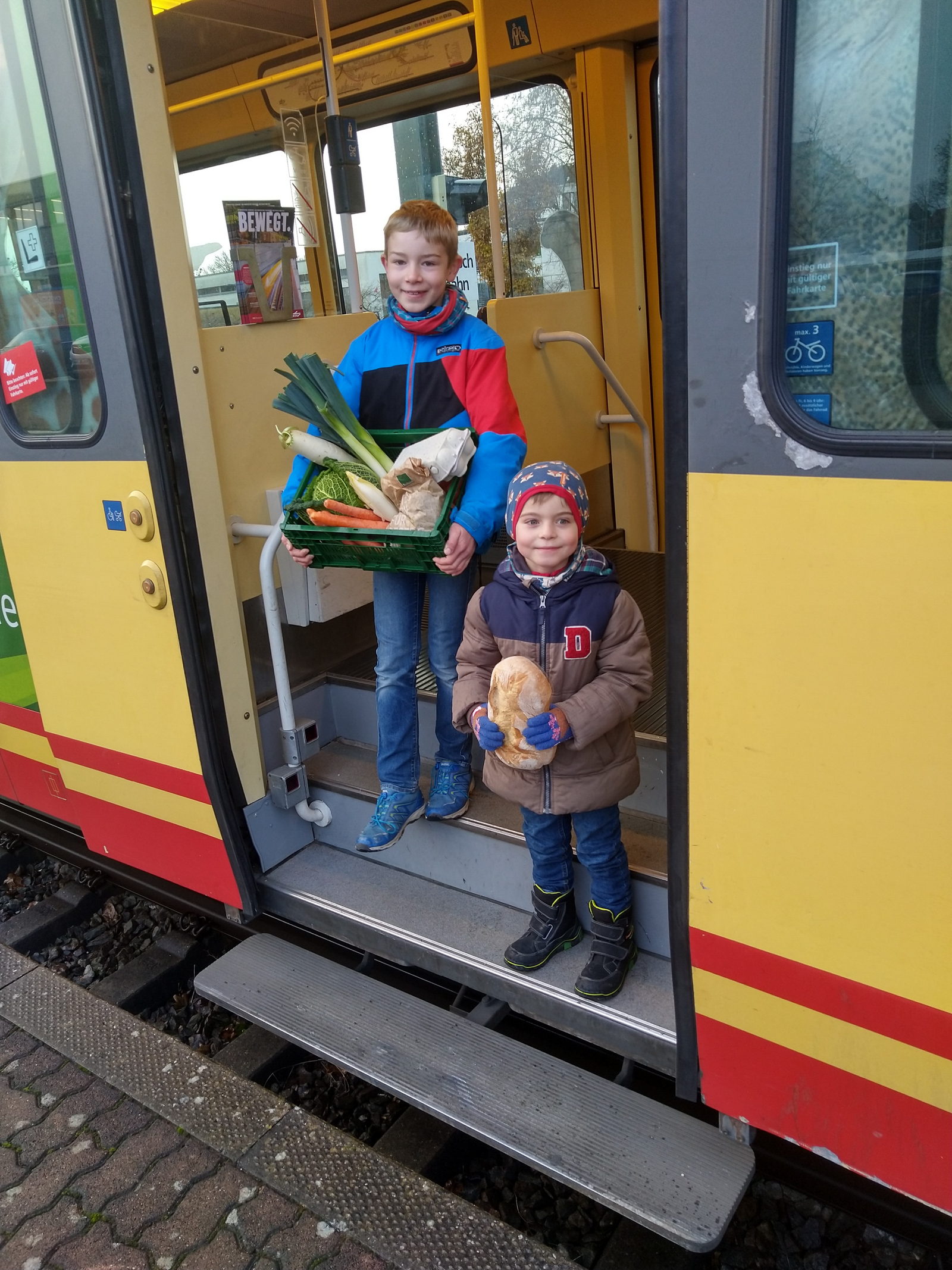 Zwei Jungen mit eine Kiste Gemüse beziehungsweise Brot in der Hand steigen aus einer Stadtbahn