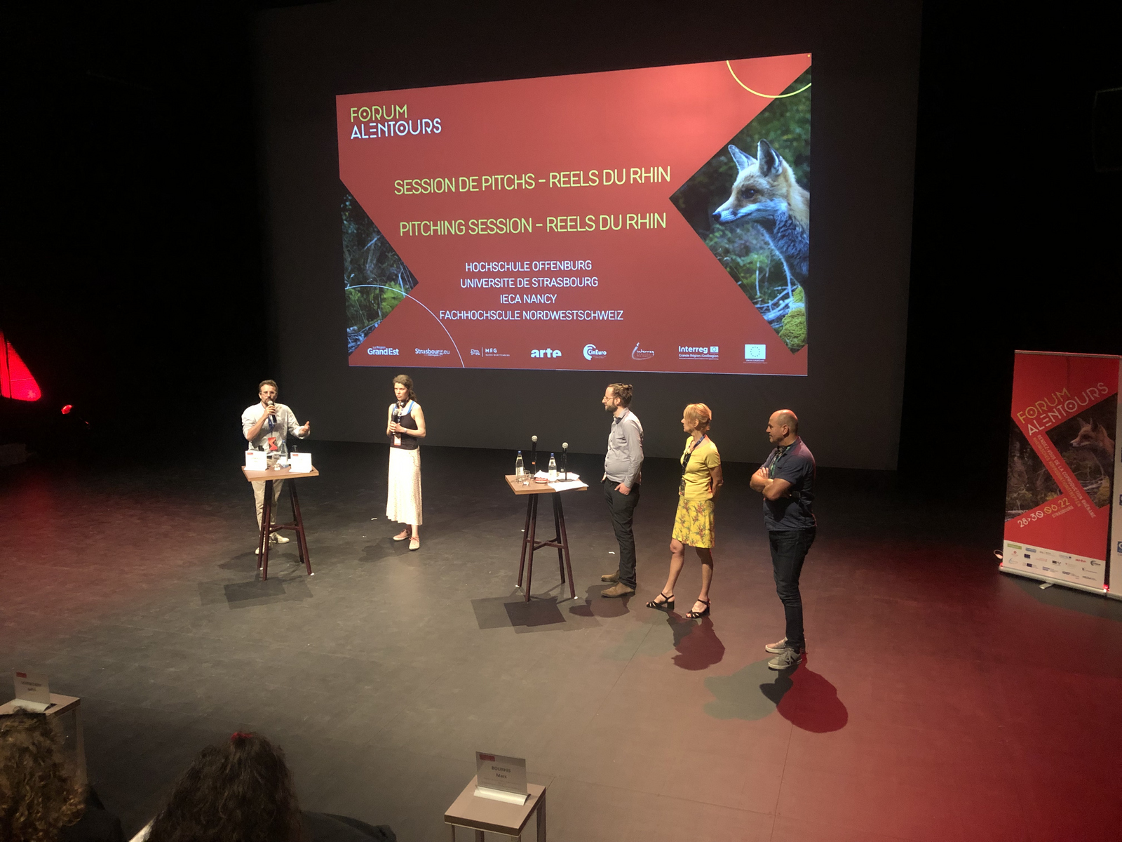 Moderierende und Jury auf der Bühne vor der Leinwand auf der die Pitches angekündigt werden